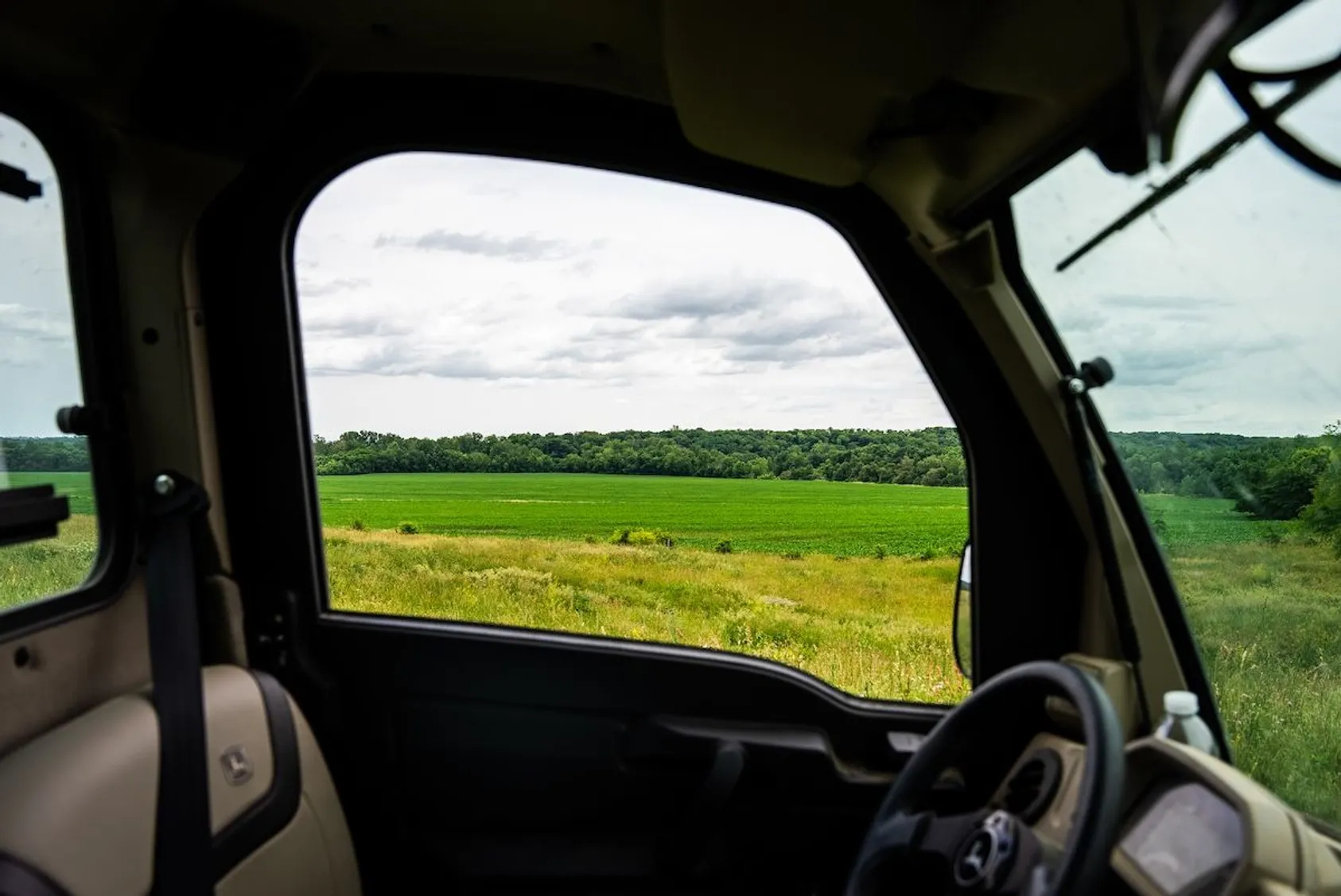 View through open vehicle tailgate showing green agricultural fields bordered by dense forest under overcast sky