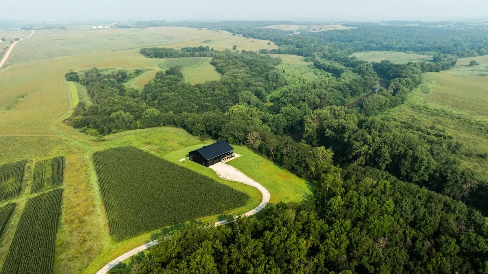 Aerial view of black barn building with gravel driveway surrounded by green crop fields and dense woodland in central Illinois