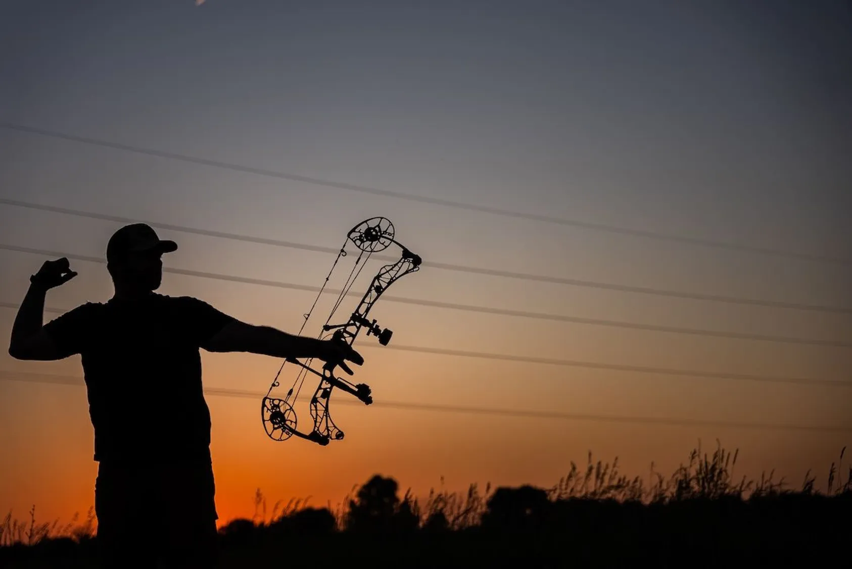 Silhouette of archer holding compound bow overhead at sunset with orange sky and tall grass in background