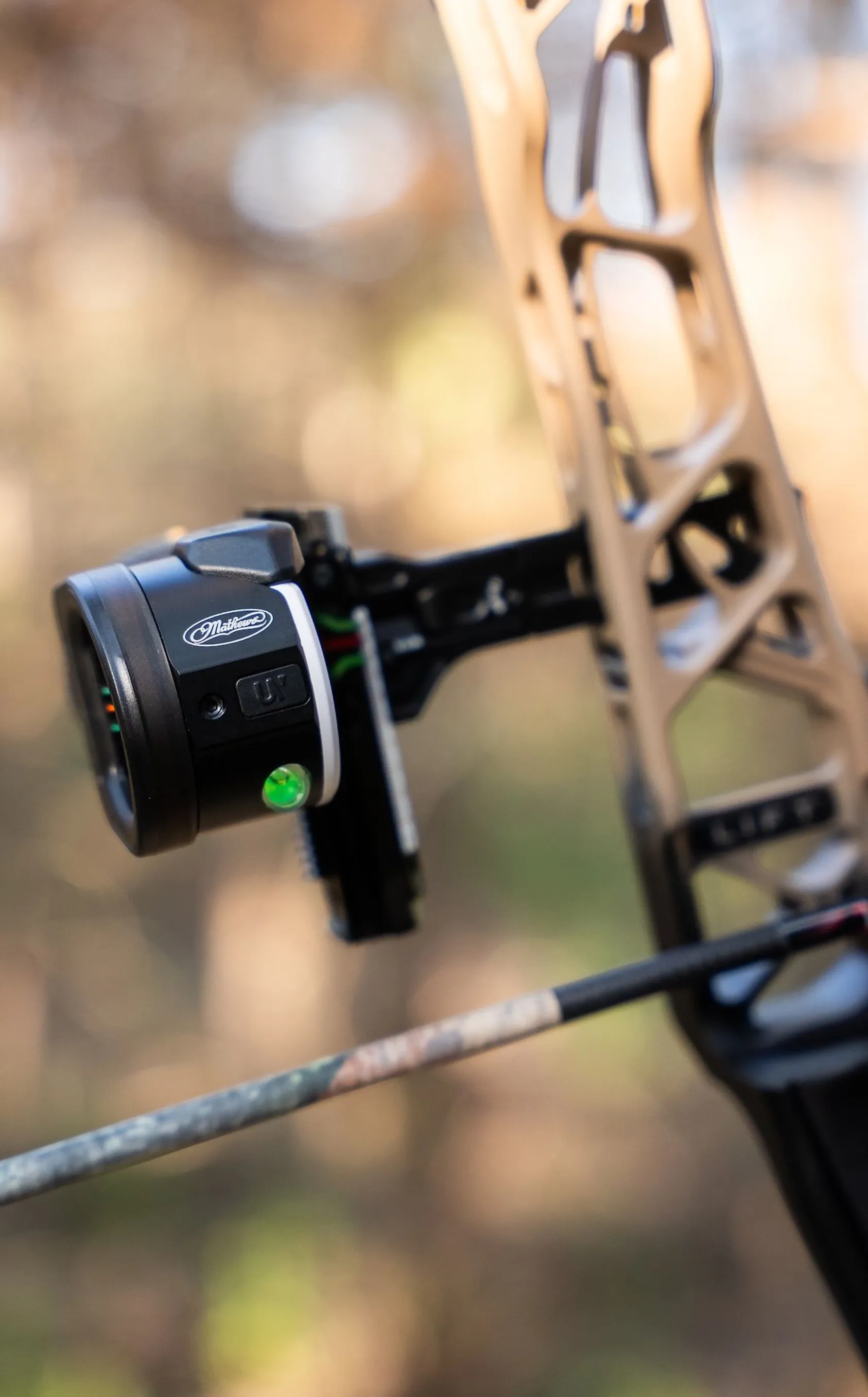 Close-up of black Mathews bow sight mounted on compound bow with green illuminated level indicator and blurred background