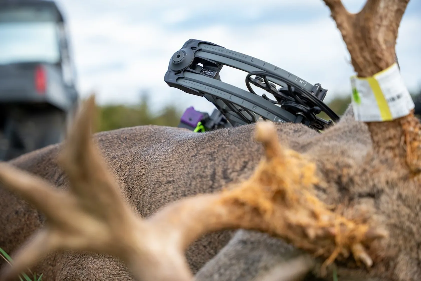Gray folding crossbow cocking device positioned on deer antlers with blurred bow and outdoor equipment in background