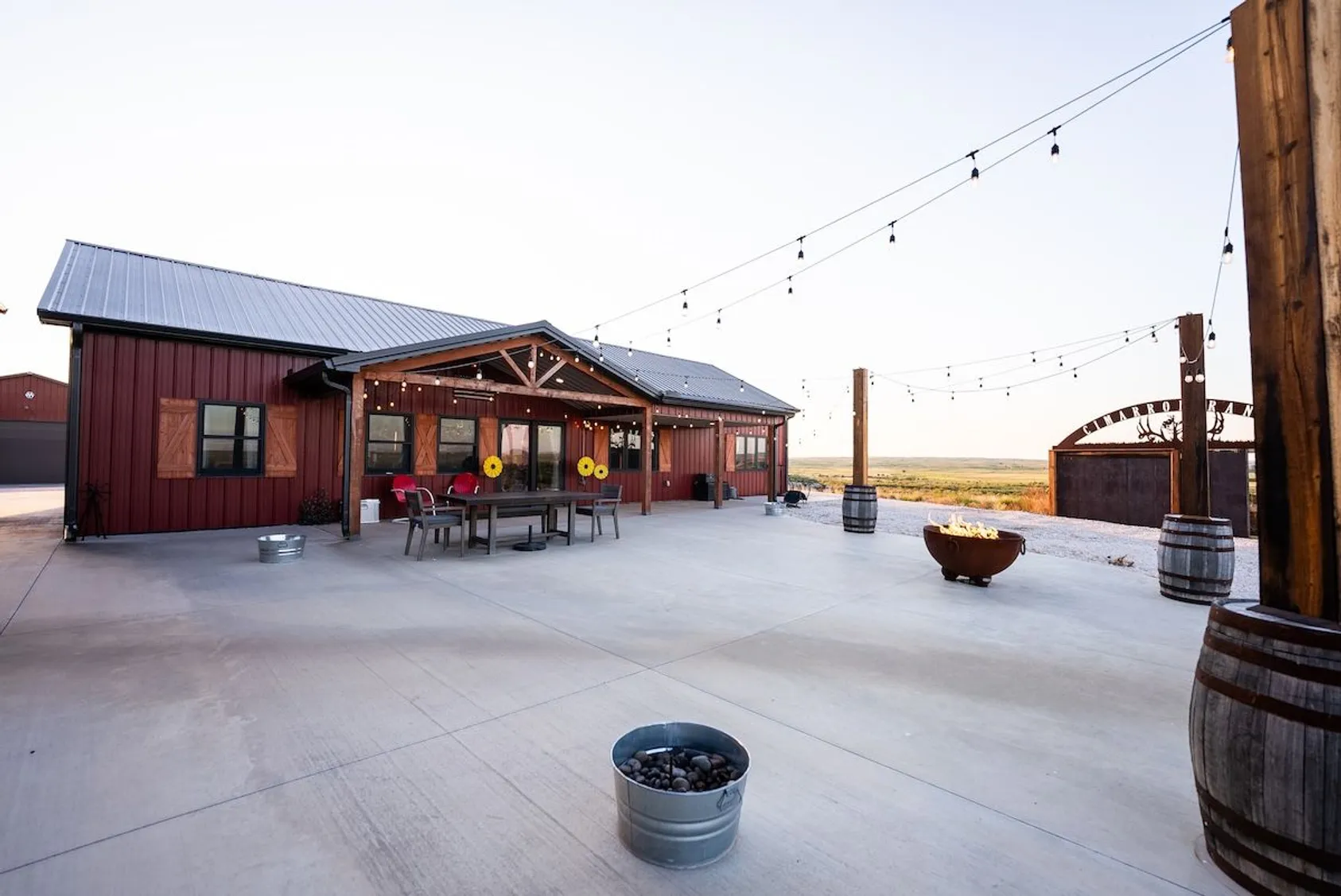 Red barn-style hunting cabin with covered patio, outdoor furniture, fire pit, and string lights on concrete patio overlooking open plains