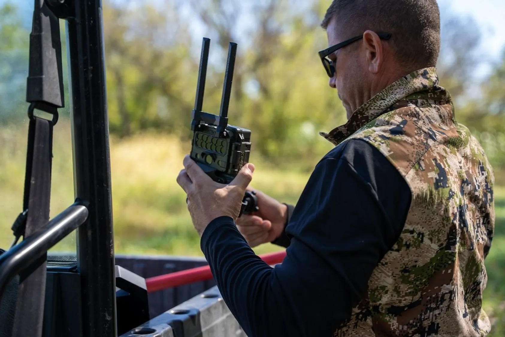 Hunter in camouflage using radio communication equipment while monitoring trail cameras from vehicle, showing modern hunting technology integration