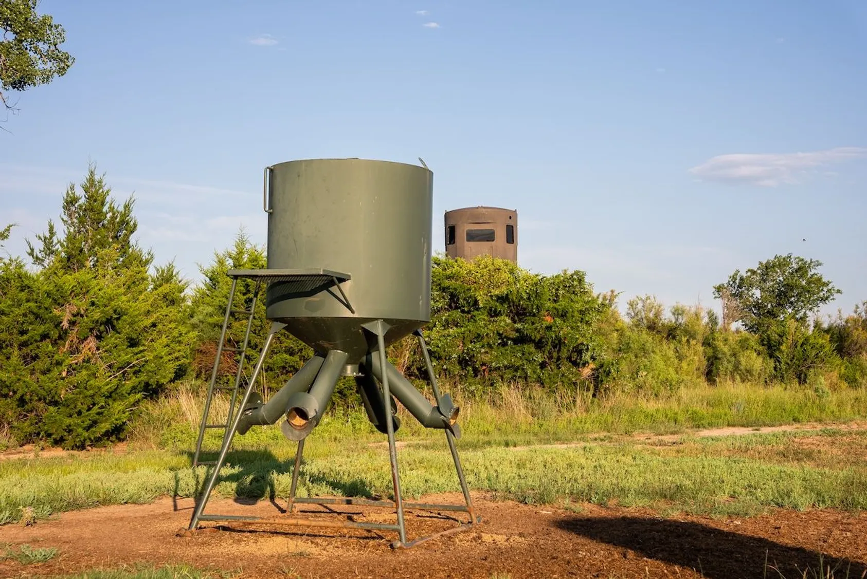 Large green cylindrical hunting blind or feeder on elevated legs in open field, showing strategic placement for wildlife attraction and monitoring