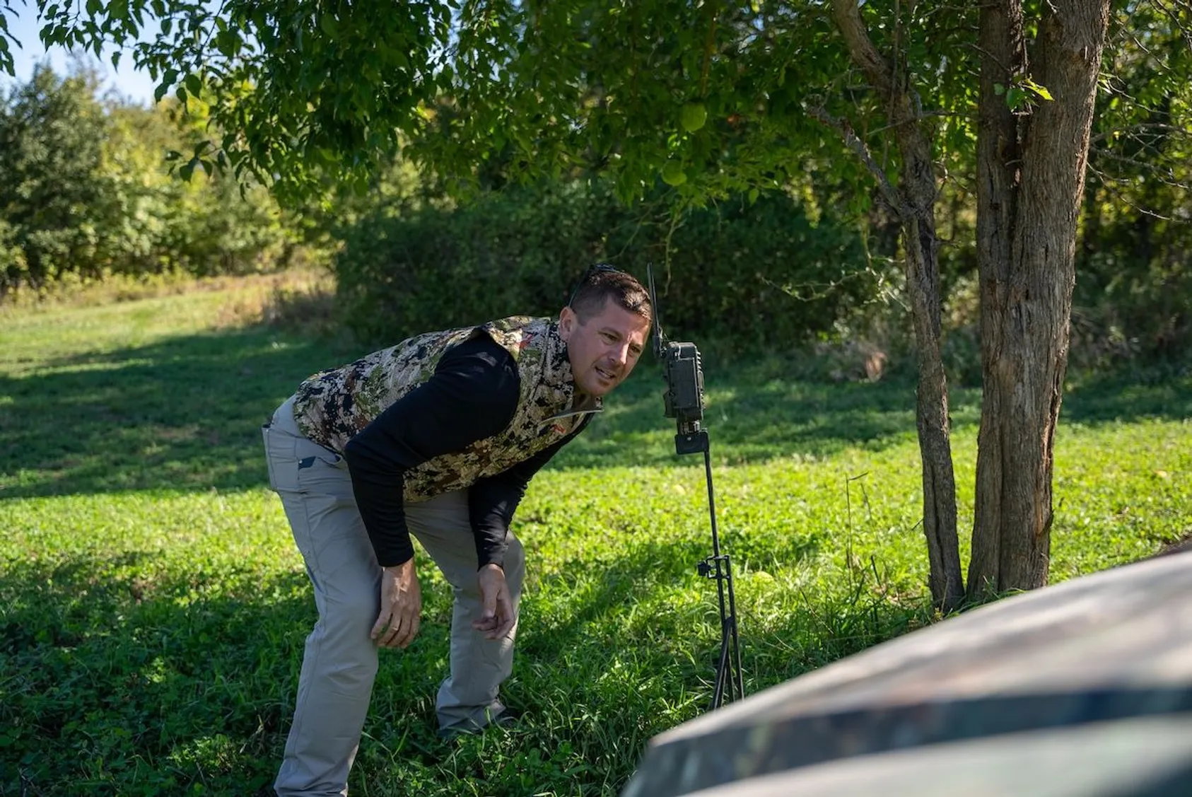 Hunter in camouflage gear checking or adjusting trail camera mounted on tree, demonstrating hands-on camera management in the field