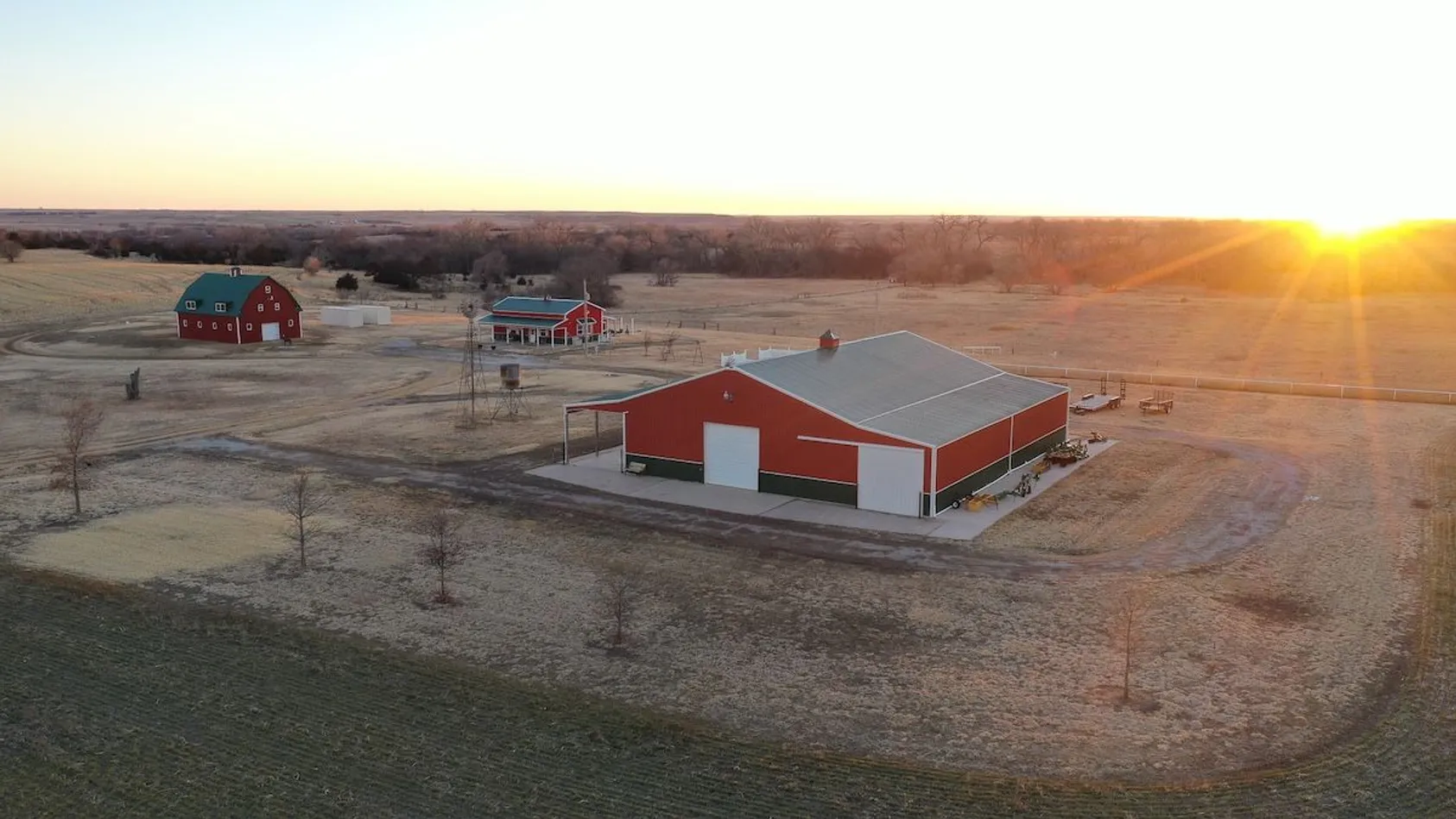 Aerial sunrise view of red barn buildings with metal roofs on farm property with golden fields and bare trees.