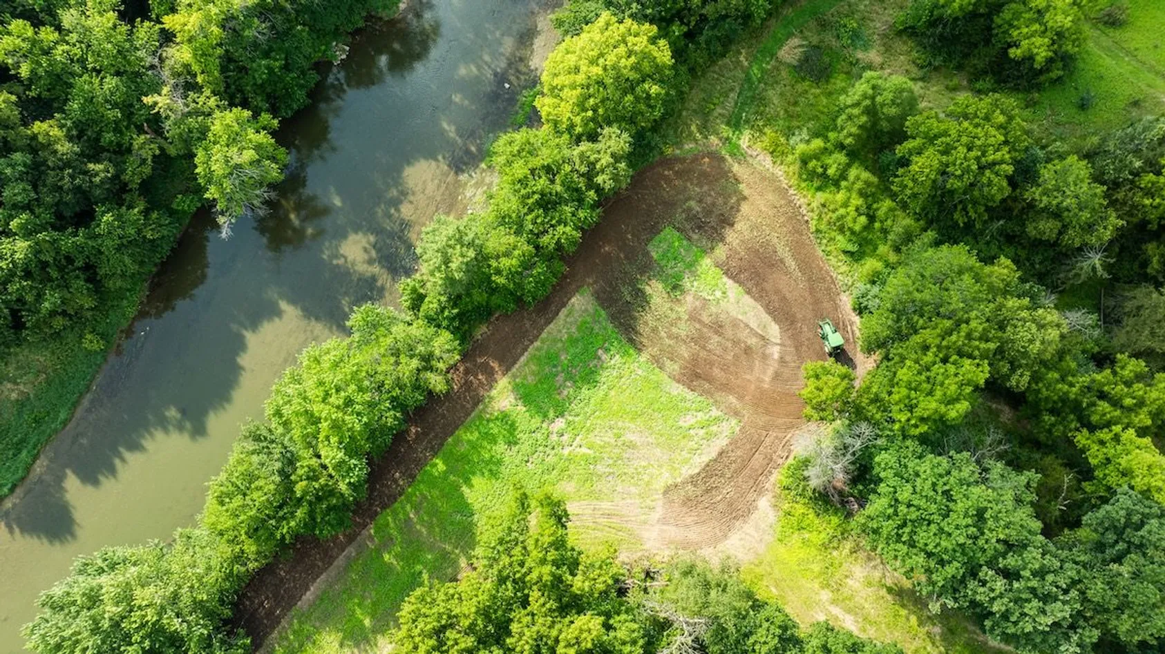 Overhead view of a tractor clearing land near a curved waterway surrounded by trees and vegetation