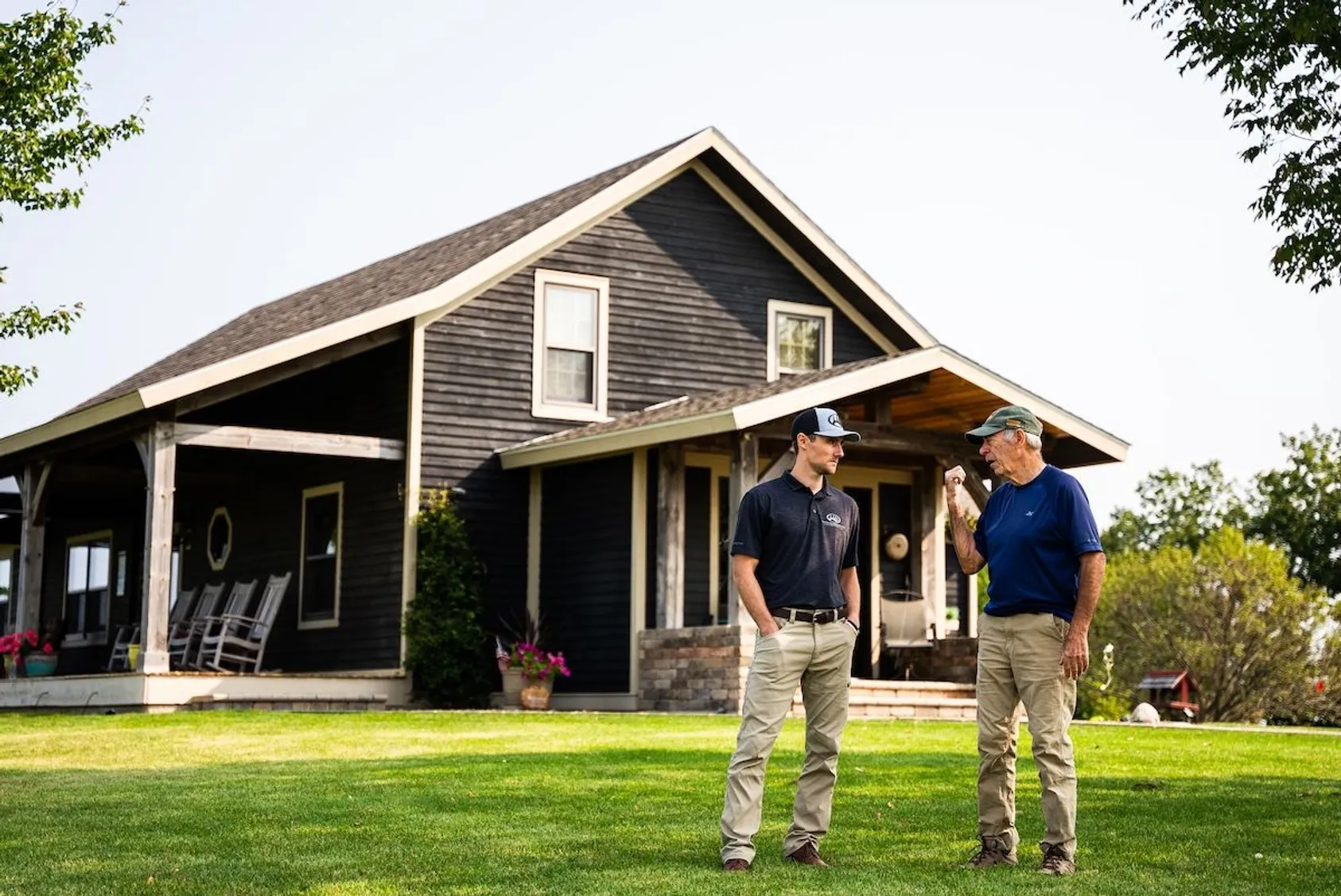 Two men in caps discussing outdoors in front of a dark gray house with a covered porch