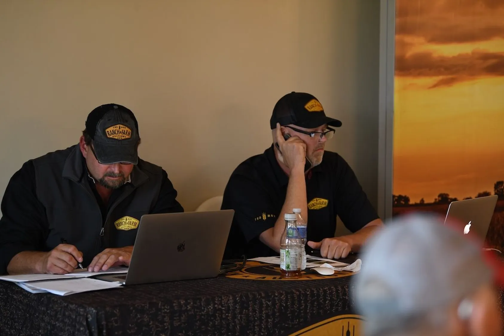 Two men in matching Ranch & Farm branded caps and shirts working on laptops at table during sunset