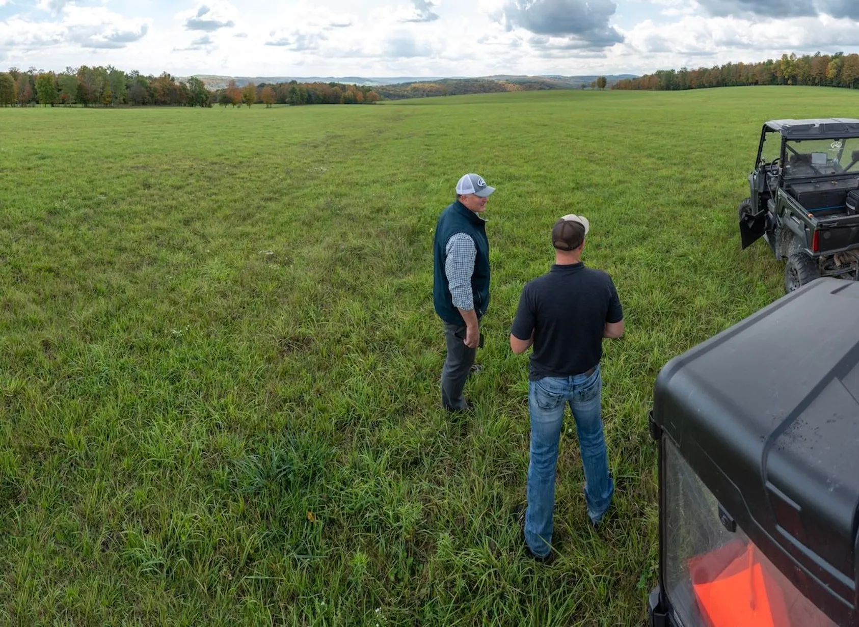 Two farmers inspecting a green pasture field with utility vehicles parked nearby, hills visible in the background.