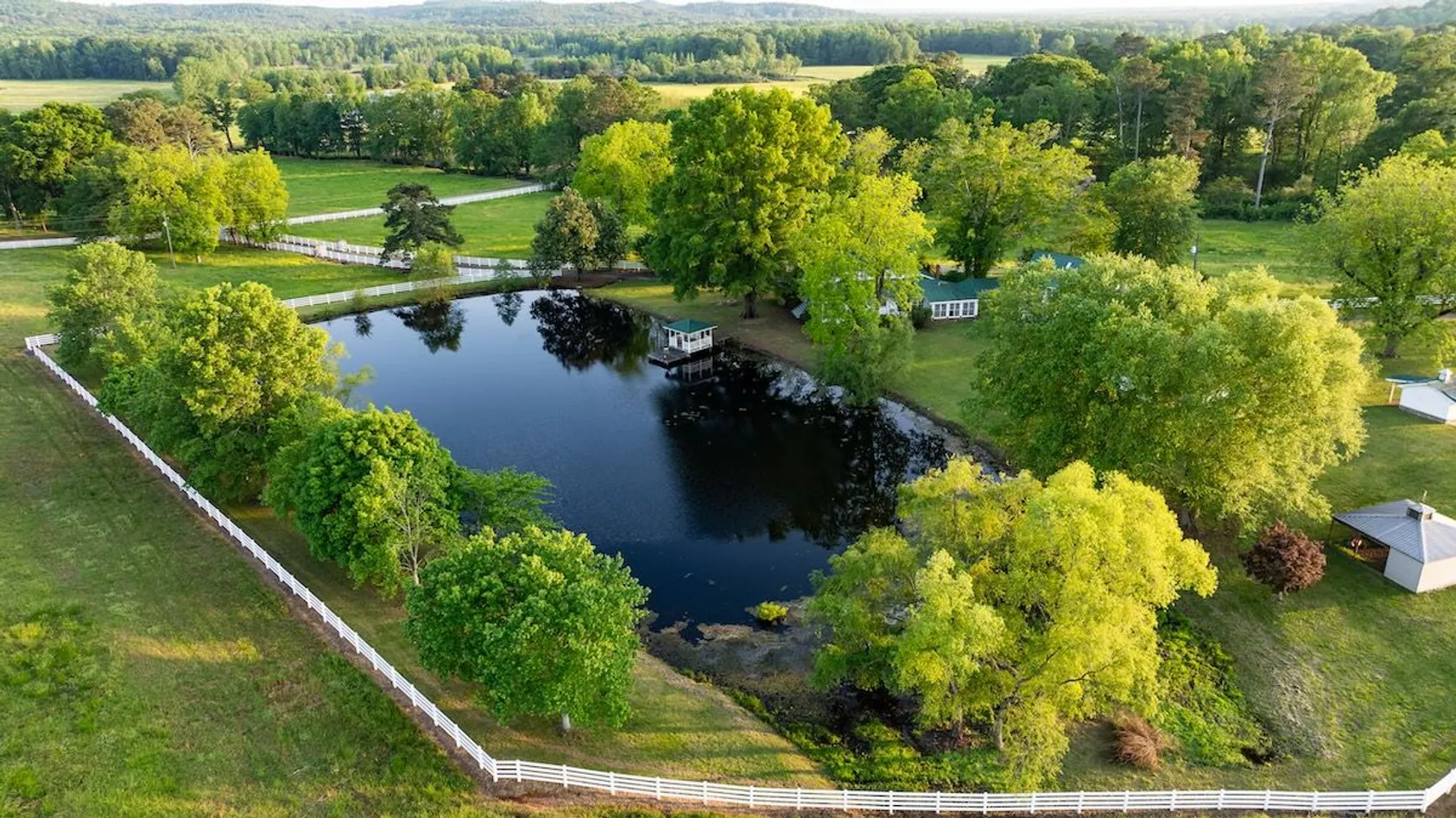 Aerial view of dark pond with white fence, dock, and buildings surrounded by bright green trees on lawn.