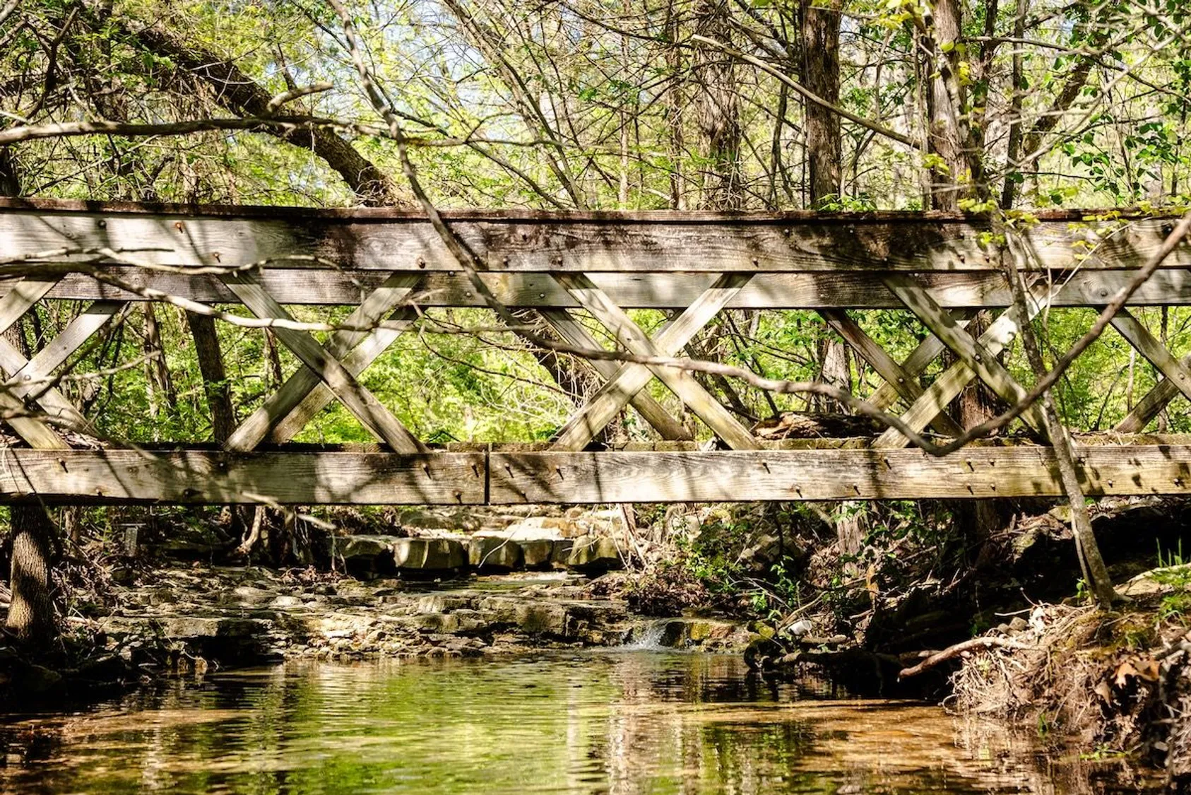 Old weathered wooden truss bridge with diagonal cross bracing spanning forest creek with moss-covered rocks and debris.