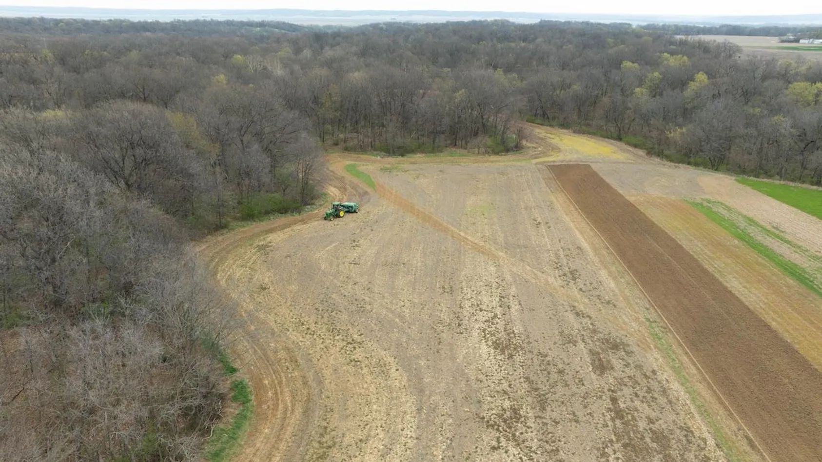 Aerial view of green tractor working in field bordered by bare winter trees with patches of varied soil conditions.