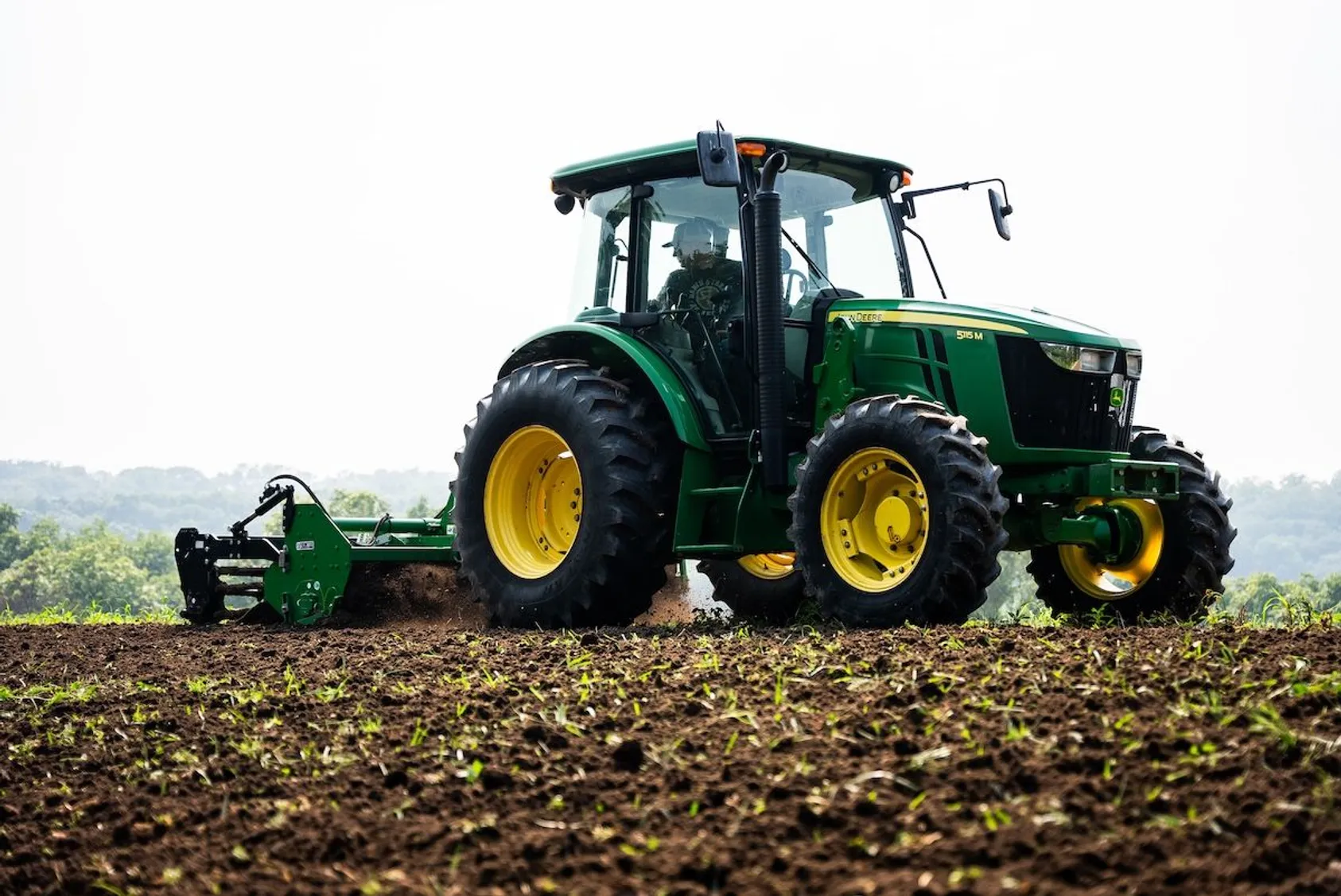 Green John Deere tractor with cultivation equipment working in field with emerging crop rows under overcast sky.