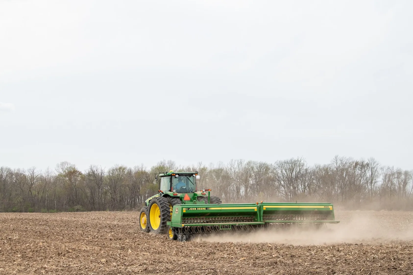 Green John Deere tractor pulling grain drill seeder across brown plowed field near bare winter trees in dusty conditions