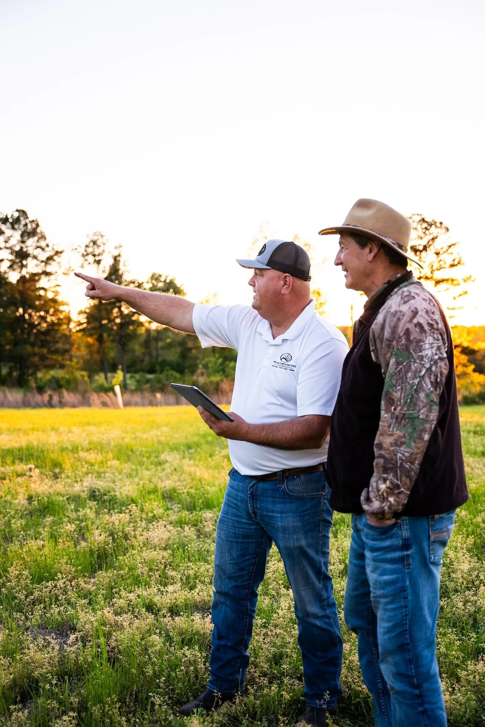 Real estate agent holding tablet and pointing across field while consulting with client in cowboy hat at sunset