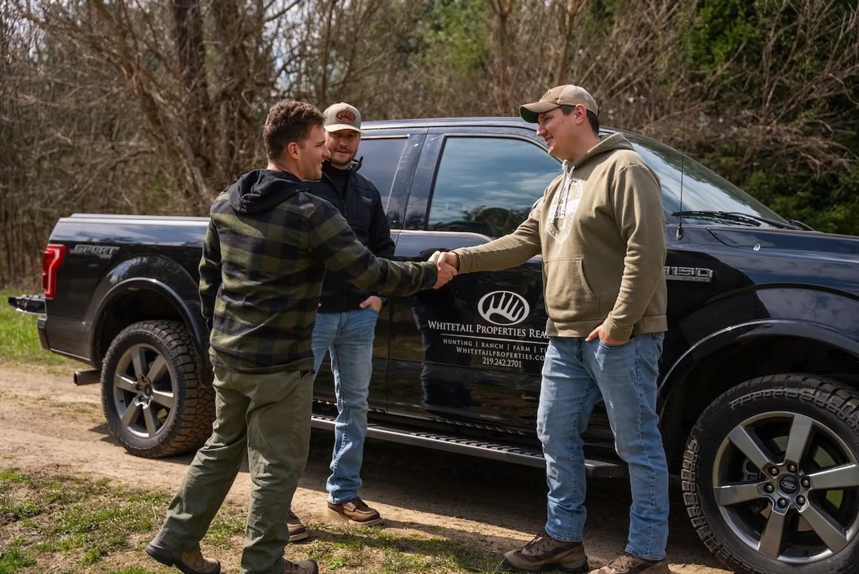 Two men shaking hands beside Whitetail Properties Real Estate truck on dirt road with bare trees in background