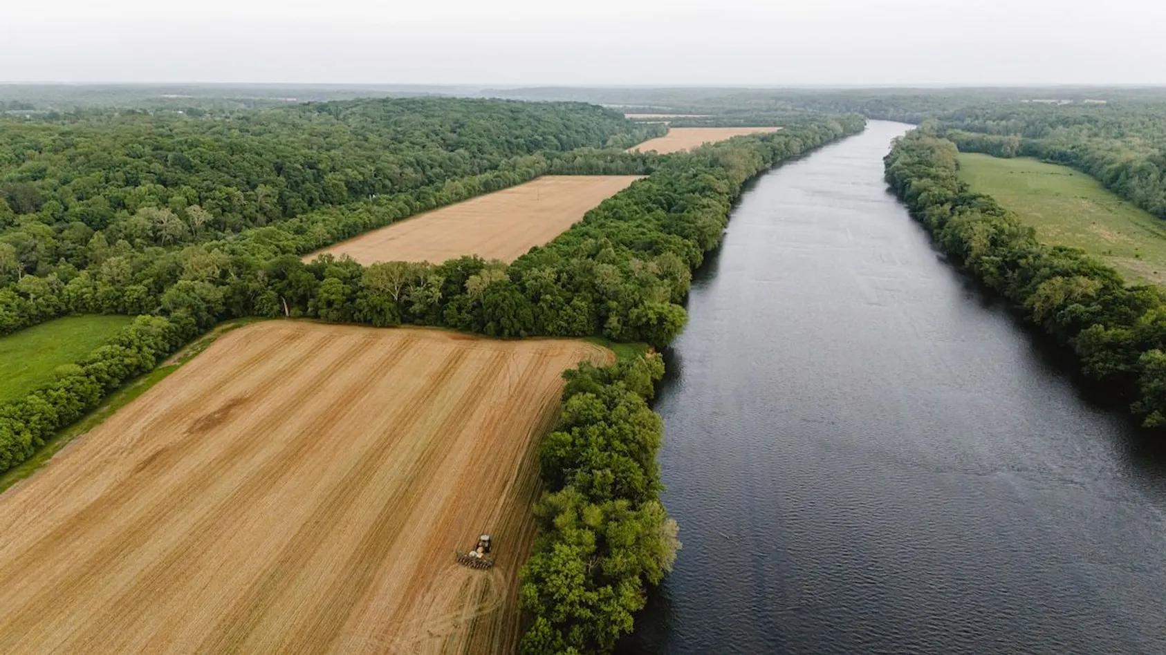 Aerial view of wide river winding between tilled brown agricultural fields and dense green forest under hazy sky with tractor working field.