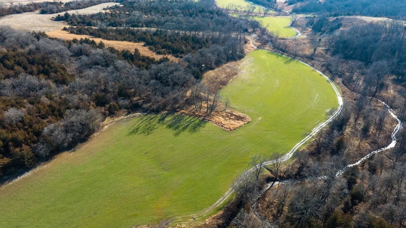 Aerial view of green pasture fields bordered by creek with white fencing and surrounded by wooded hills.