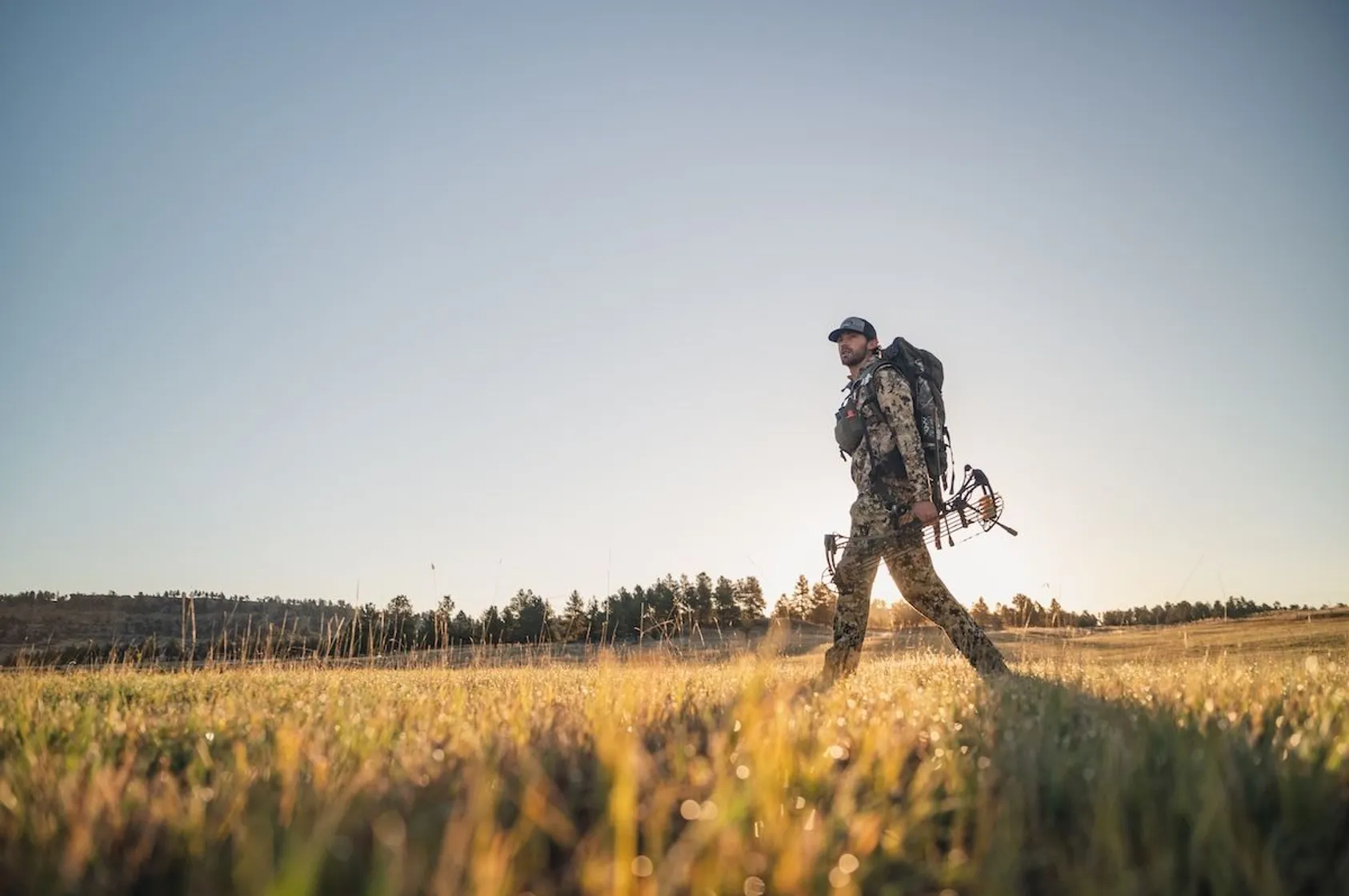 Bowhunter in camouflage with backpack and compound bow walking through golden grassland at sunrise.