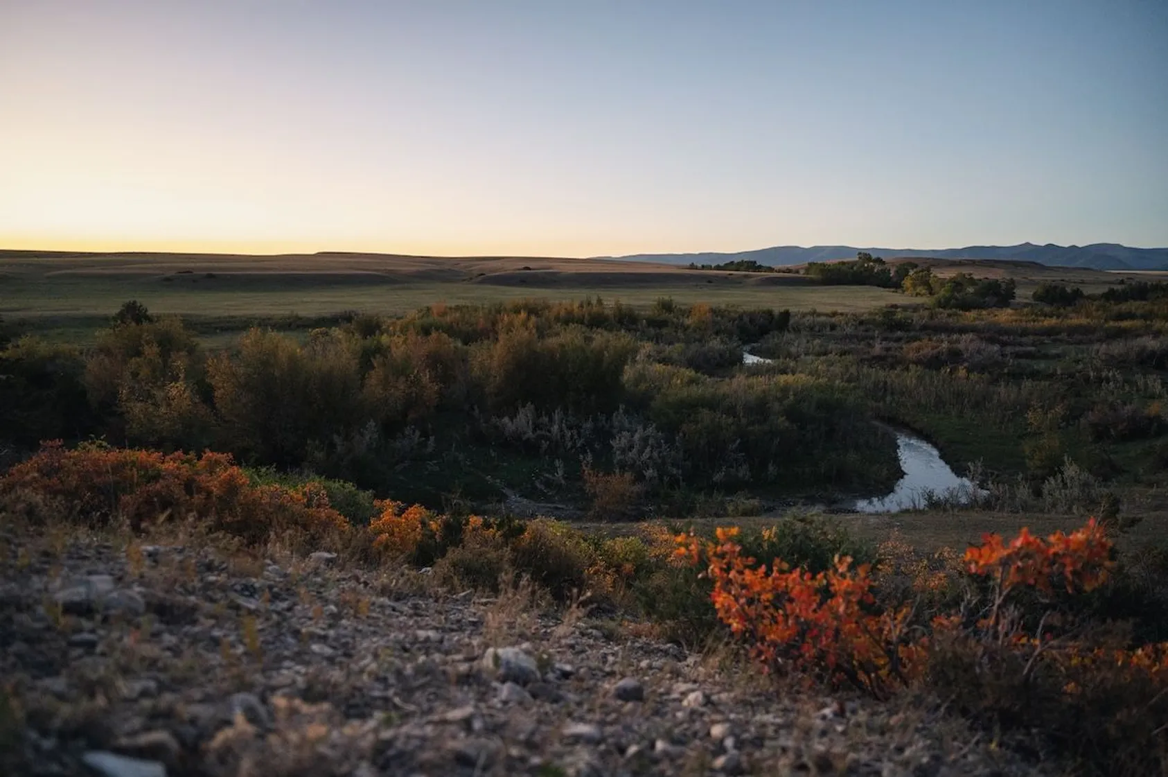 Sunset view over western prairie landscape with winding creek through autumn foliage and distant mountains.