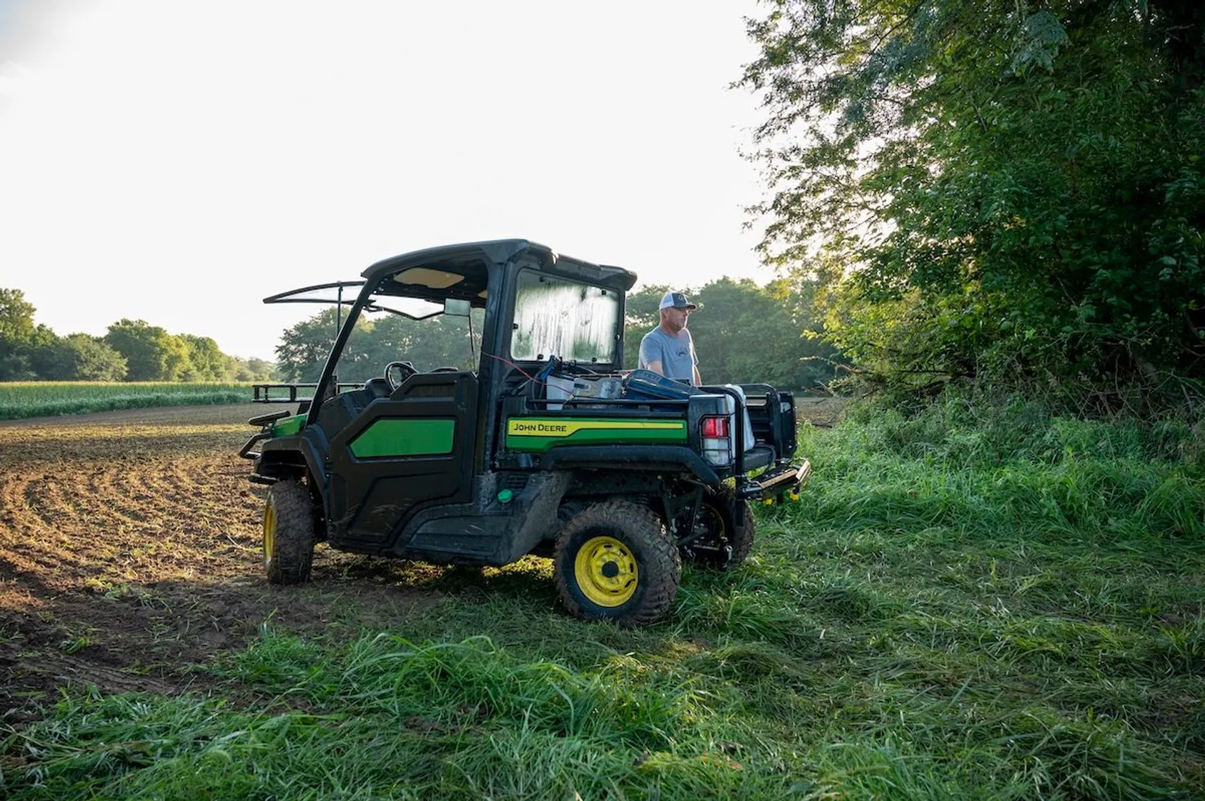 Person standing beside John Deere utility vehicle at edge of tilled field with green vegetation and trees.