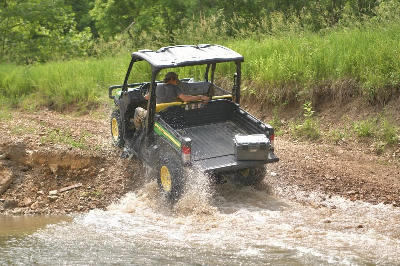 John Deere utility vehicle with storage box splashing through muddy water puddle on dirt trail.