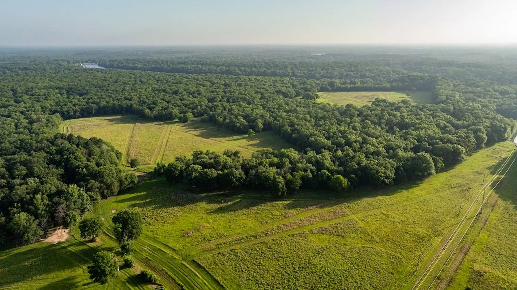 Aerial view of wooded landscape with green fields and power lines visible at right edge in hazy conditions.