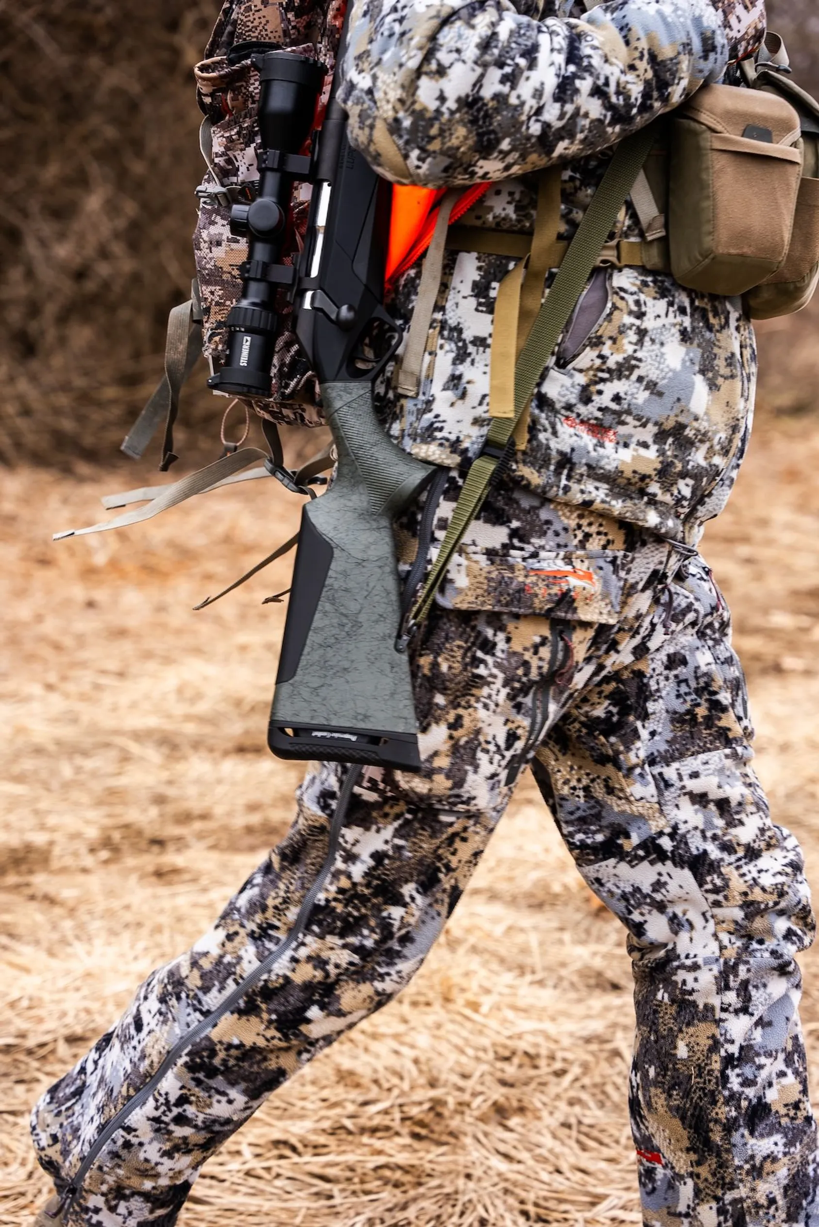Hunter in digital camouflage clothing carrying rifle with sling across back walking through dry grass and brush