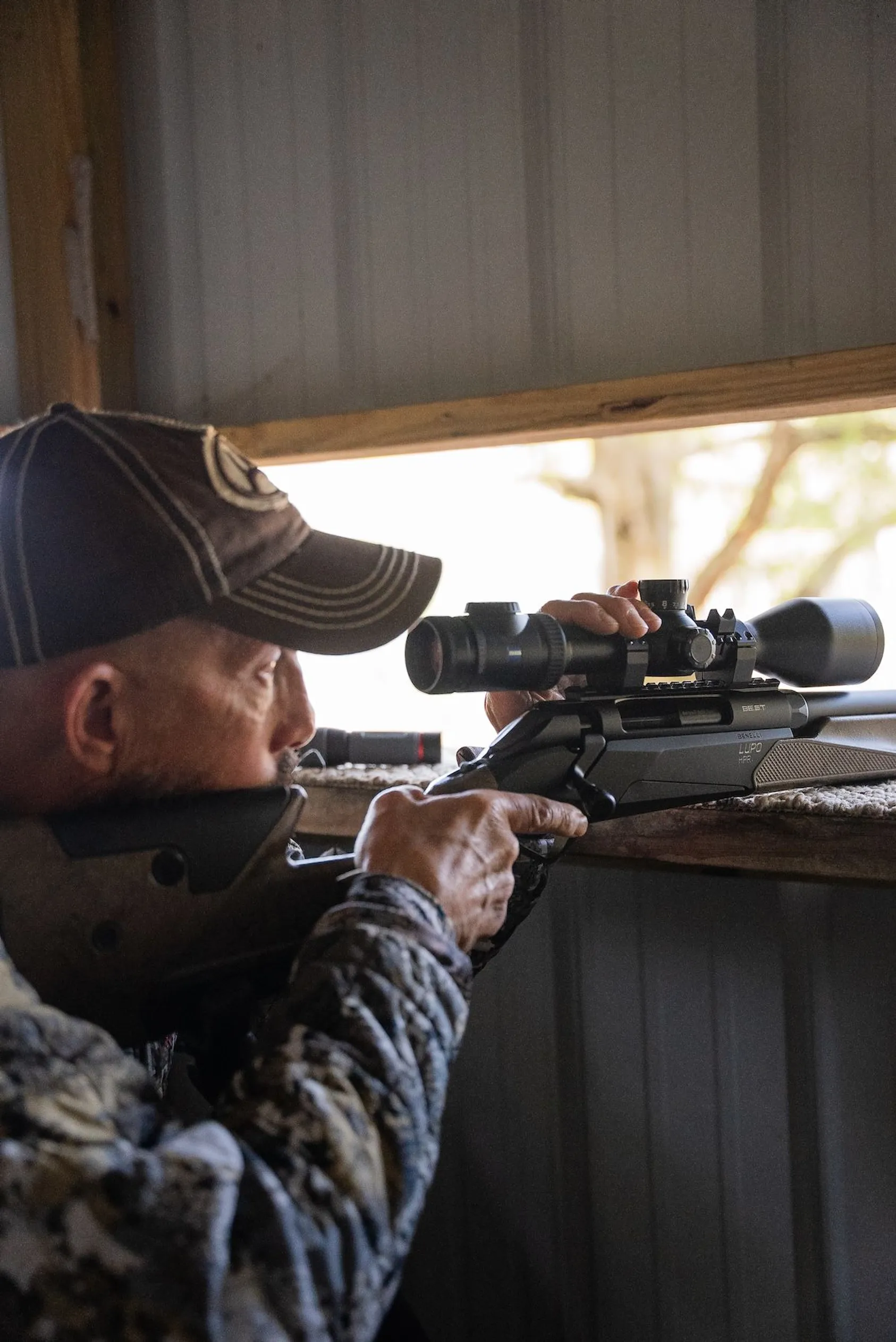 Hunter in cap and jacket aiming scoped rifle while positioned at wooden shooting rest inside hunting blind
