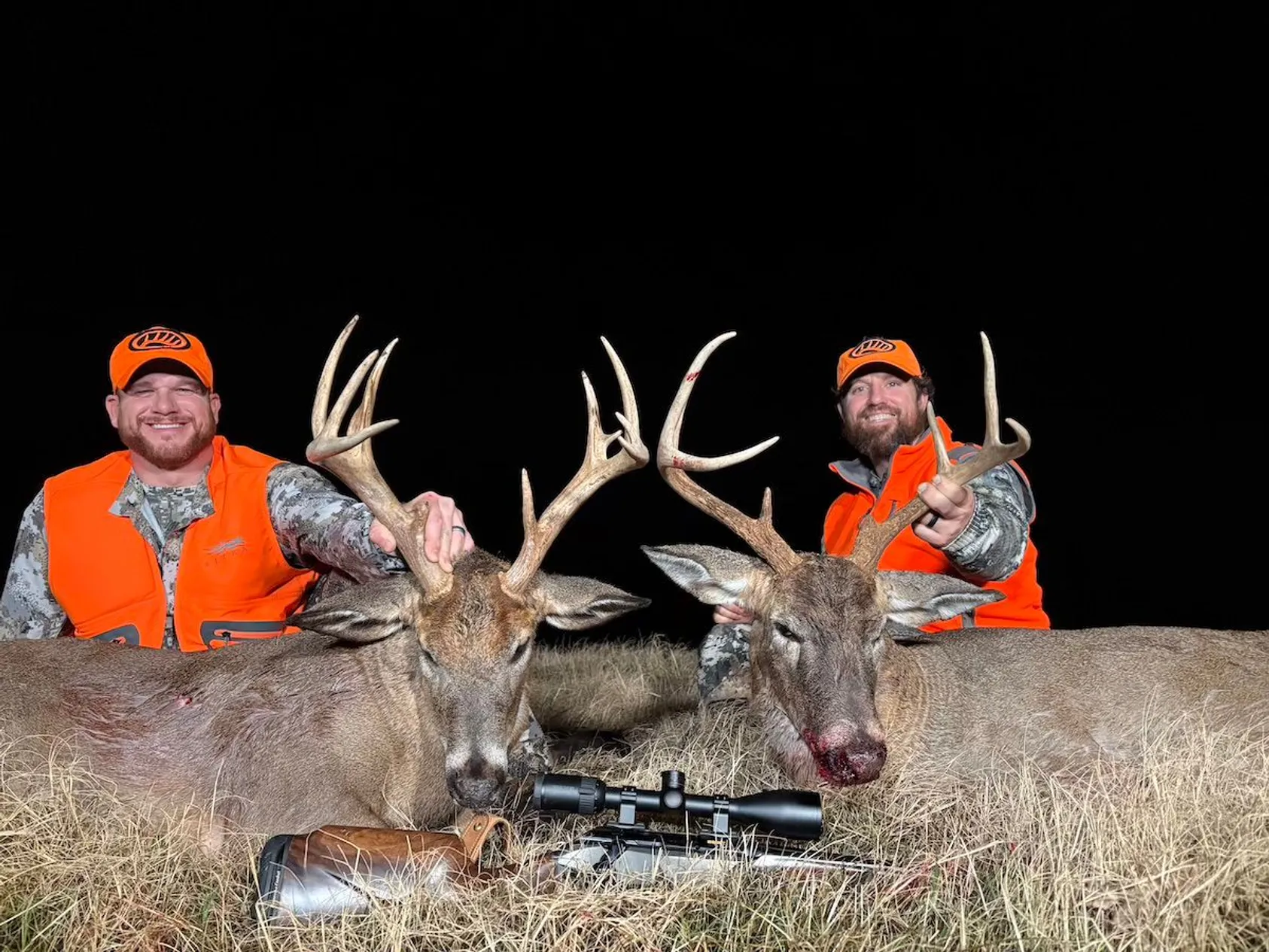 Two hunters in orange safety vests and caps posing behind two harvested white-tailed bucks with large antlers and scoped rifle